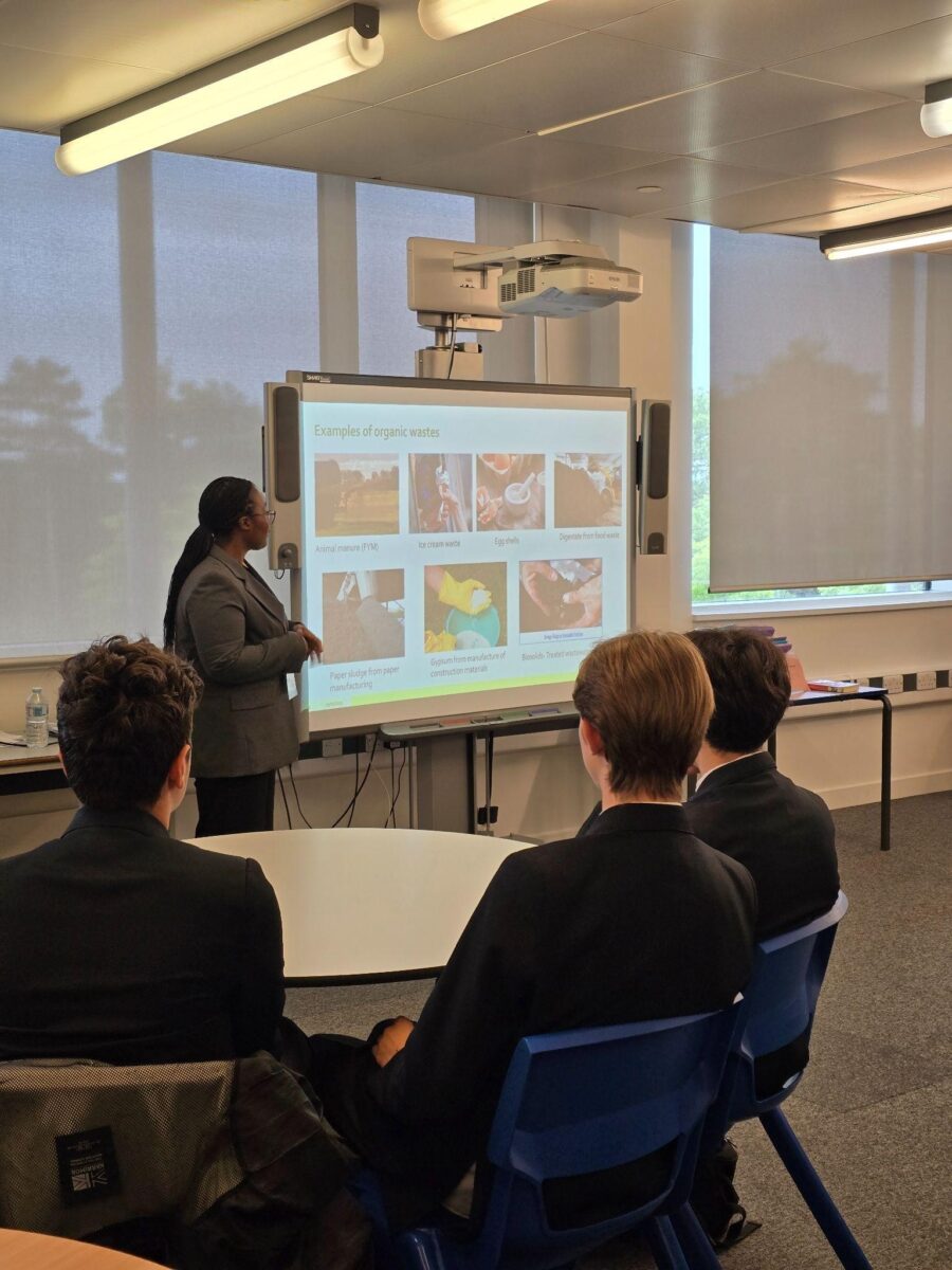 A classroom setting shows a presenter standing to the left of an interactive whiteboard displaying the title "Examples of organic wastes" and several images illustrating different types of organic waste. The presenter, facing the whiteboard, appears to be addressing a group of students seated in the foreground. The students are wearing dark blazers and are looking towards the presenter and the screen. The classroom is well-lit with fluorescent lighting and features large windows with blinds in the background. A projector is mounted on the ceiling, directed at the whiteboard. The overall scene depicts a lesson or presentation taking place in a modern classroom.
