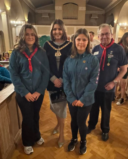 A group of four individuals is captured in a medium shot, standing indoors on a polished wooden floor. From left to right, the group consists of Elsie, a woman wearing a dark blue jacket, black trousers, and white trainers, with a red neckerchief tied around her neck. Next to her stands a woman with shoulder-length brown hair, adorned with a ceremonial chain with a pendant, a dark blazer, and a light grey skirt. To her right is Chloe, sporting a teal jacket, black leggings, and black trainers. Completing the group is a man on the far right, wearing a dark blue polo shirt, dark trousers, and black shoes, with a red patterned scarf tied around his neck. All four are looking towards the camera with smiles. The background features a room with light-colored walls, arched architectural details, and warm lighting from overhead fixtures.