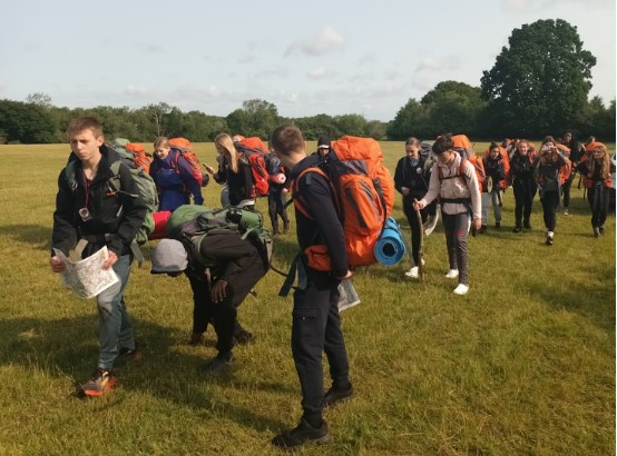 The image shows a large group of young people, likely teenagers, walking across a wide, open grassy field under a bright sky. Almost everyone in the group is carrying a large backpack, many of which are brightly colored (orange, blue, grey). Several individuals are also holding maps, suggesting they are navigating during an expedition or hike. The attire is casual and practical for outdoor activity. In the background, there are trees lining the edge of the field. The overall scene strongly indicates a Duke of Edinburgh (DofE) expedition or a similar outdoor challenge where participants carry their own gear and navigate.