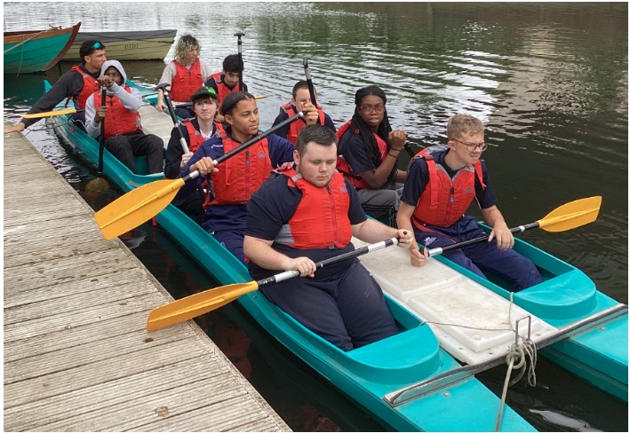 This picture shows a group of people, appearing to be mostly young adults or teenagers, in several boats on the water, next to a wooden dock. In the foreground, there are two teal-colored boats that look like multi-person canoes or kayaks, possibly connected together. Most of the people in these boats are wearing red life vests and holding yellow paddles. Some people are looking towards the camera, while others are looking forward or to the side. In the background, there are other boats docked, including a wooden rowboat. The water appears calm, and there's some greenery or trees visible in the distance on the opposite bank.