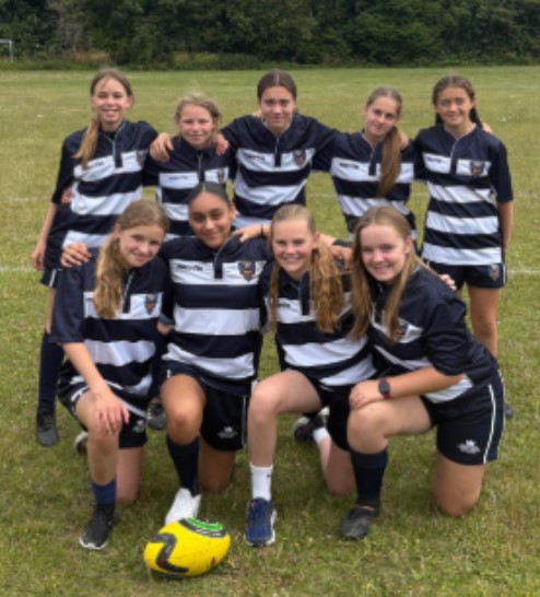 The image displays a group of nine young females, likely a sports team, posing on a grassy field. They are all wearing matching navy blue and white striped sports jerseys and dark shorts, which suggests a team uniform, possibly for rugby given the "Rugby7s.jpg" filename. A yellow and green rugby ball is visible in the foreground. Some of the girls are standing, while others are kneeling in front, forming a group photo. They are all smiling, indicating a positive and cohesive team spirit. The background shows more grassy area and some trees.