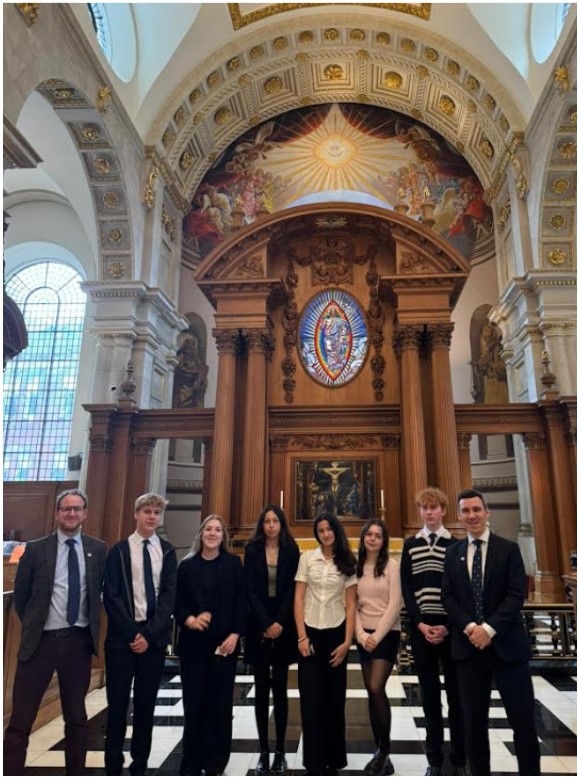 This picture shows a group of people, two adults and six young people, standing inside a grand church. The church features a large, ornate wooden structure with an altarpiece, and a highly decorated ceiling with a large mural. There are also stained-glass windows visible on the left side of the image.