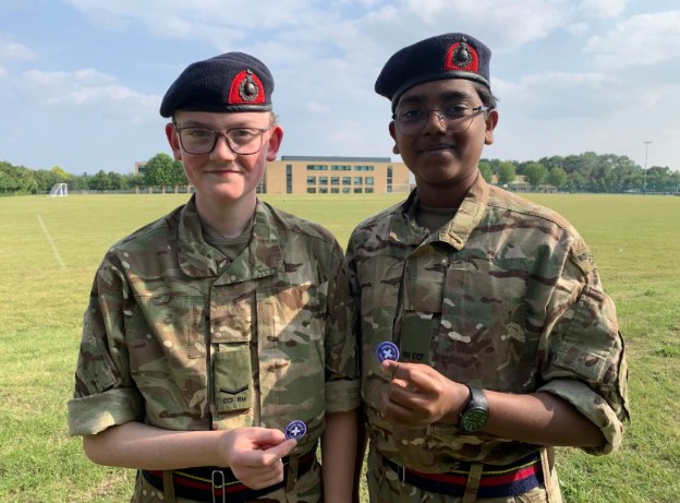 The image features two young people in what appear to be military-style camouflage uniforms and berets, standing on a grassy field. Both are smiling at the camera and holding up small circular badges or coins. The person on the left is wearing glasses. The person on the right is also wearing glasses and a watch. In the background, there's a modern building and open green fields under a clear sky. Their attire and the badges suggest they might be part of a cadet program or similar youth organization.
