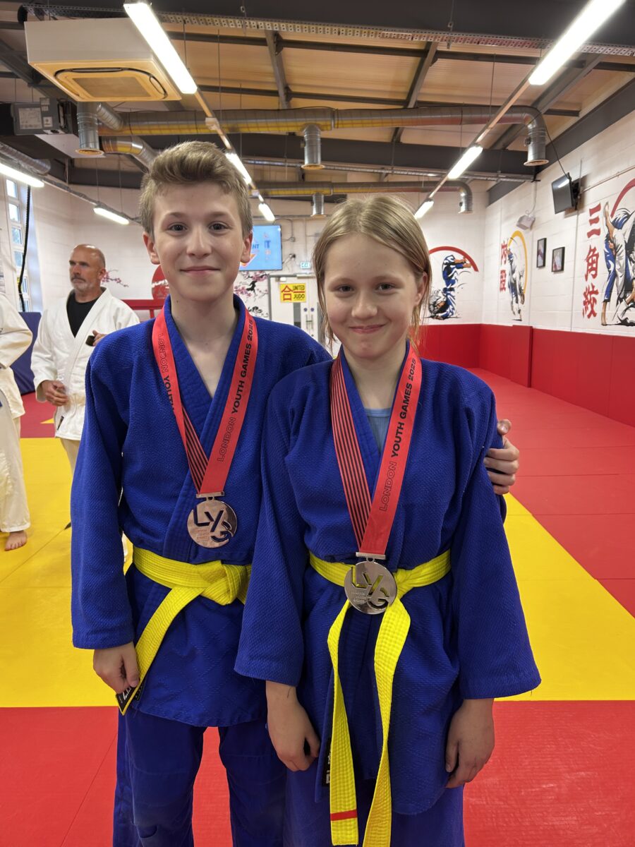 he image shows two young individuals, a boy and a girl, standing side-by-side and smiling at the camera. Both are wearing blue judogi (judo uniforms) with yellow belts, indicating their rank in martial arts. Each has a medal with a red ribbon around their neck, which reads "LONDON YOUTH GAMES 2023", suggesting they have participated in and achieved success at this event. They appear to be in a dojo or training hall, with red and yellow mats on the floor and martial arts-related decor on the walls in the background. An adult in a white judogi is partially visible in the left background. The overall impression is one of achievement and camaraderie after a competition.