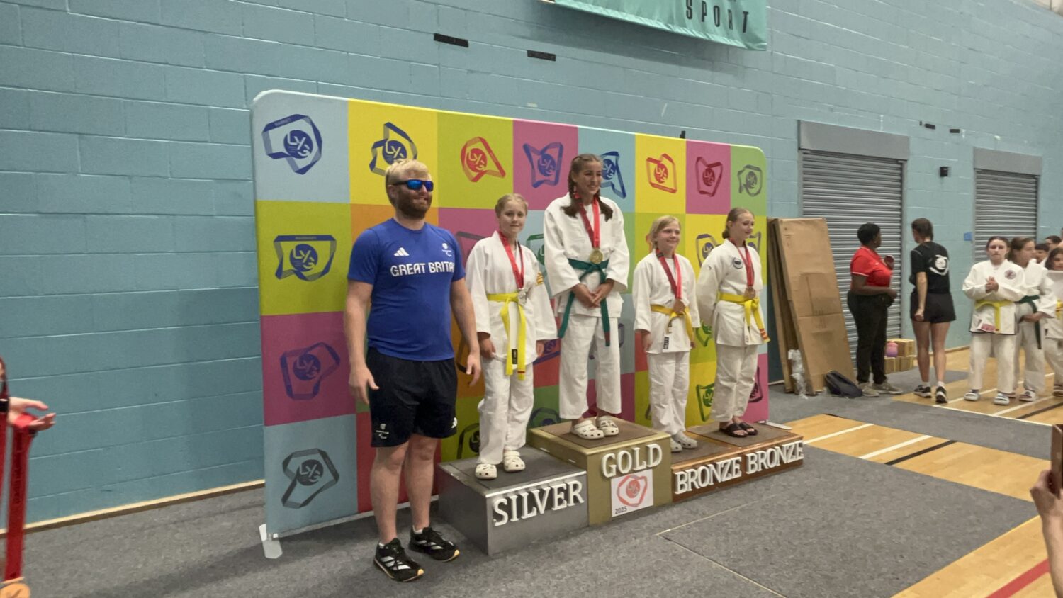 The image captures a medal ceremony, likely for a judo or martial arts competition, in an indoor setting. On a multi-tiered podium (labeled "GOLD", "SILVER", "BRONZE"), four young girls in white judogi (martial arts uniforms) with various colored belts (yellow and green) are standing. The girl on the gold podium is wearing a medal around her neck. To the left of the podium, a man in a blue "GREAT BRITAIN" t-shirt and sunglasses is standing, seemingly an instructor or coach, next to the "SILVER" position. The background features a colorful wall with various logos or symbols. On the right, other participants in judogi are visible, observing the ceremony. The scene suggests a formal recognition of achievement in a sporting event.