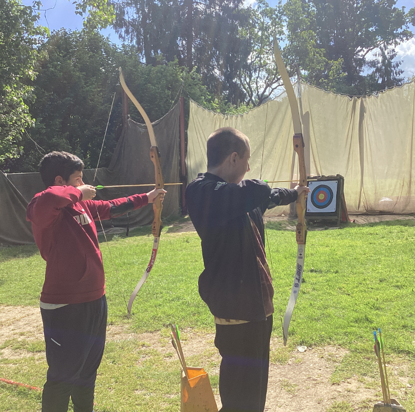 The image shows two young individuals participating in archery outdoors. Both are holding recurve bows and appear to be aiming at targets. The person on the left is wearing a maroon hoodie and is seen drawing the bow. The person on the right is wearing a dark jacket and is also in the process of aiming. In the background, there's a traditional bullseye archery target set up on a grassy area, with a net behind it for safety. The setting is clearly outdoors, with trees and sunlight, suggesting a recreational or training session at an archery range.