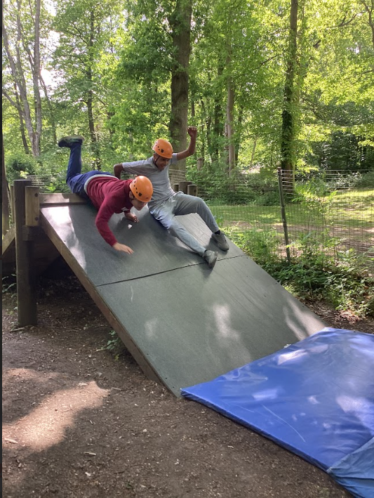 The image captures an outdoor adventure activity where two individuals are sliding down a sloped surface. They are both wearing orange helmets, indicating a safety measure for the activity. One person is lower down the slope, lying on their front, while the other is higher up, sitting and leaning back, with one arm raised. At the bottom of the ramp, there's a blue mat, likely for a soft landing. The setting is a wooded area with tall trees, suggesting a ropes course or adventure park environment. This activity appears to be a type of slide or 'zip' activity often found in adventure centers.