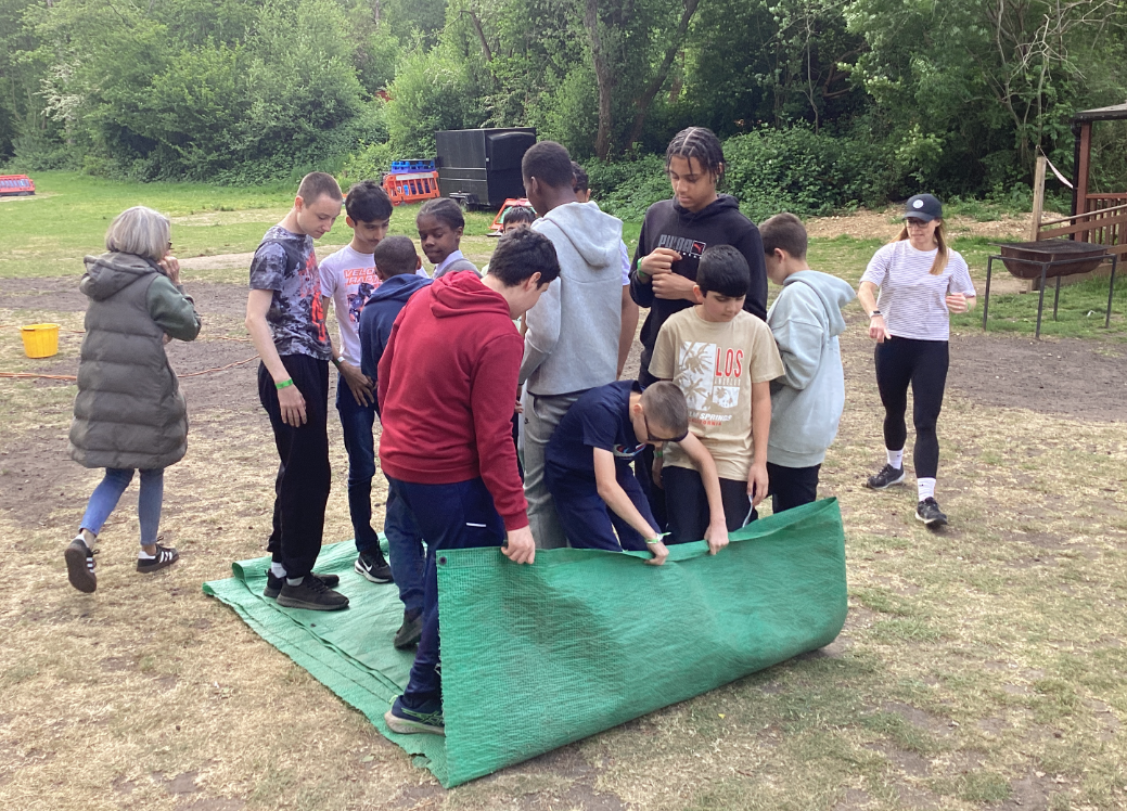 The image shows a group of young people and a couple of adults participating in an outdoor activity on a grassy area. They are gathered around a large green mat, which some of the young people are actively holding, rolling, or moving. This suggests a team-building exercise or a game that involves manipulating the mat. The participants are dressed casually. In the background, there are trees, some outdoor equipment, and what looks like a small wooden structure or shelter. The overall scene indicates an organized outdoor education or leisure activity.