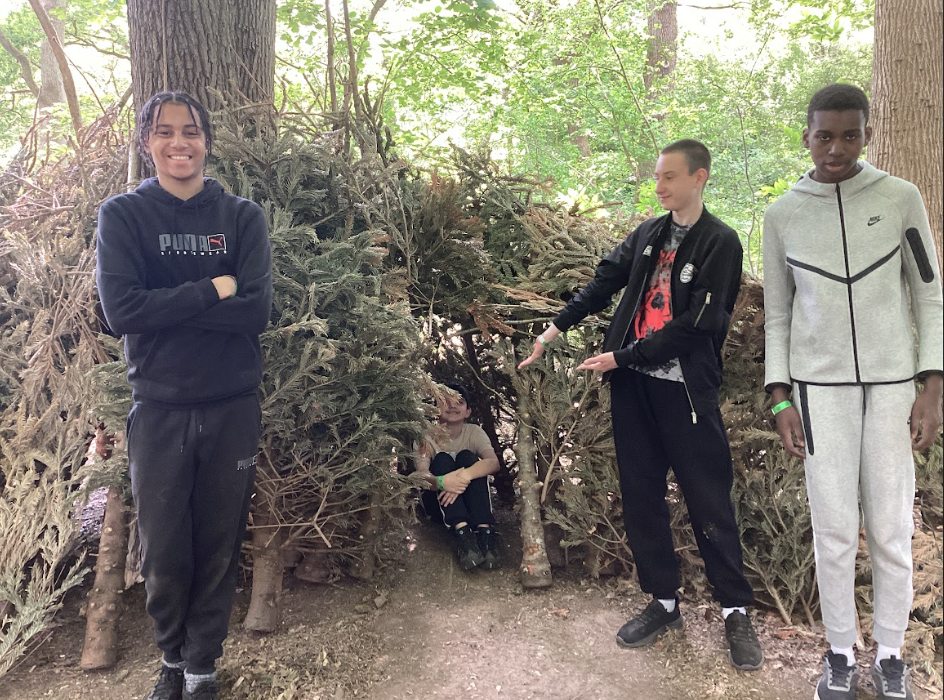 This image shows a group of four young people in a woodland setting, standing around a structure made of branches and foliage, resembling a survival shelter or den. On the left, one boy is standing with his arms crossed, smiling at the camera. He is wearing a dark hoodie and track pants. In the center, partially inside the shelter, another boy is sitting on the ground. To the right of the shelter, a boy in a dark jacket is pointing towards the shelter and smiling. On the far right, another boy is standing, looking towards the camera, wearing a light grey tracksuit. The overall scene suggests a fun outdoor activity, possibly involving bushcraft or shelter building in a natural environment.