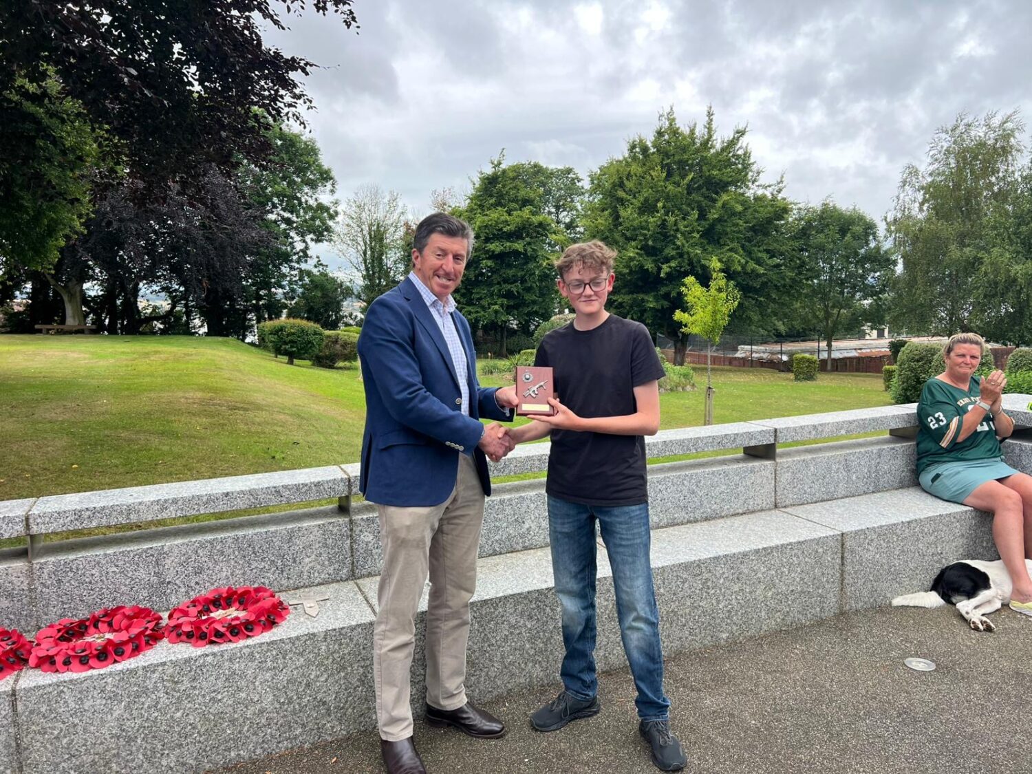 An adult male in a suit jacket is shaking hands with a younger male in a t-shirt and jeans, who is holding a small plaque or award. They are outdoors near a low stone wall with poppy wreaths on it. Another person is sitting on the wall in the background.