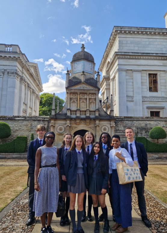 A group of students in school uniforms, accompanied by an adult, are standing outside in front of a grand architectural building on a sunny day.