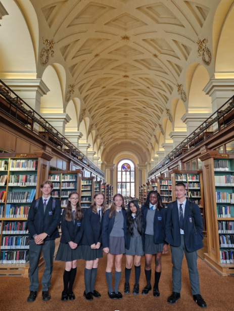A group of students in school uniforms are standing in what appears to be a large library or hall with tall bookshelves and an ornate, arched ceiling.