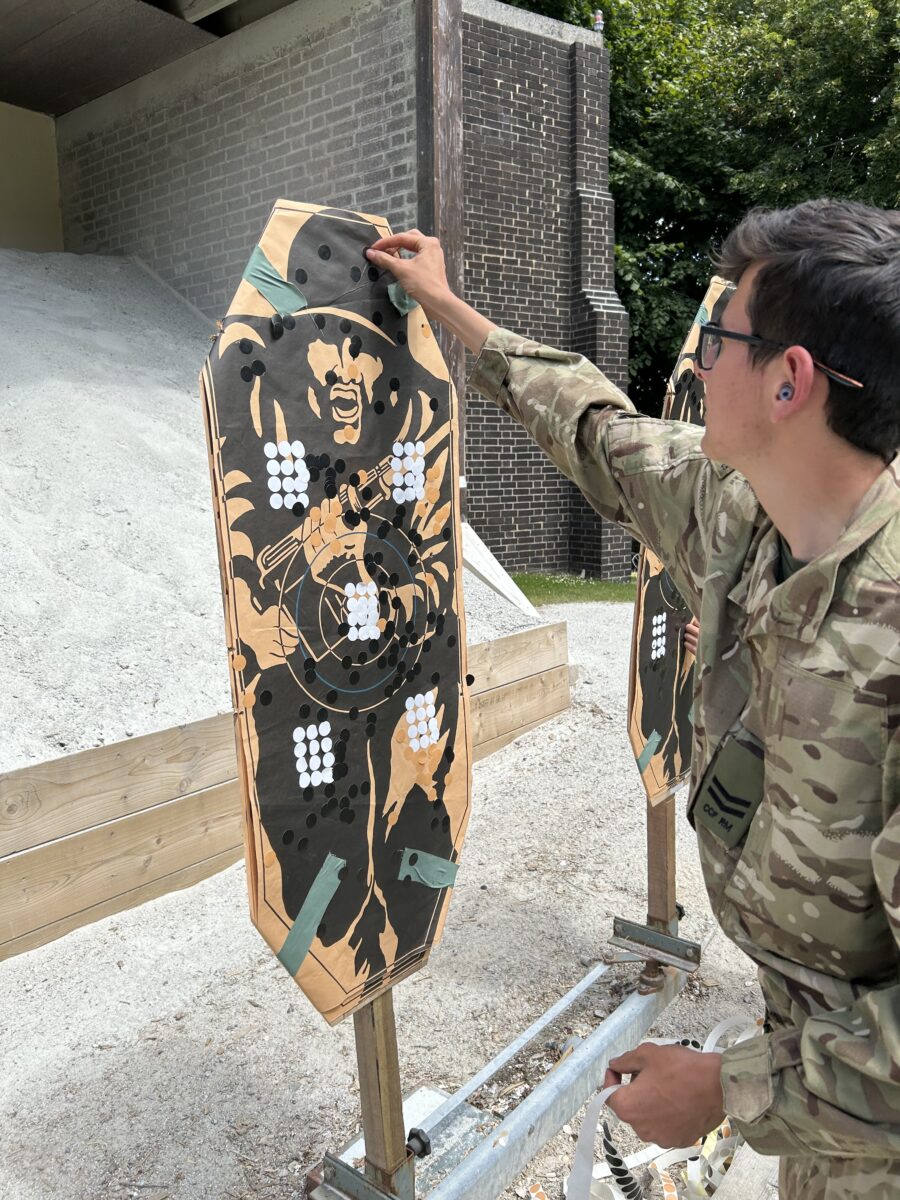 A person in camouflage fatigues with safety glasses and ear protection is standing at a shooting range, affixing or adjusting a target. The target has several white patches indicating where shots have landed.
