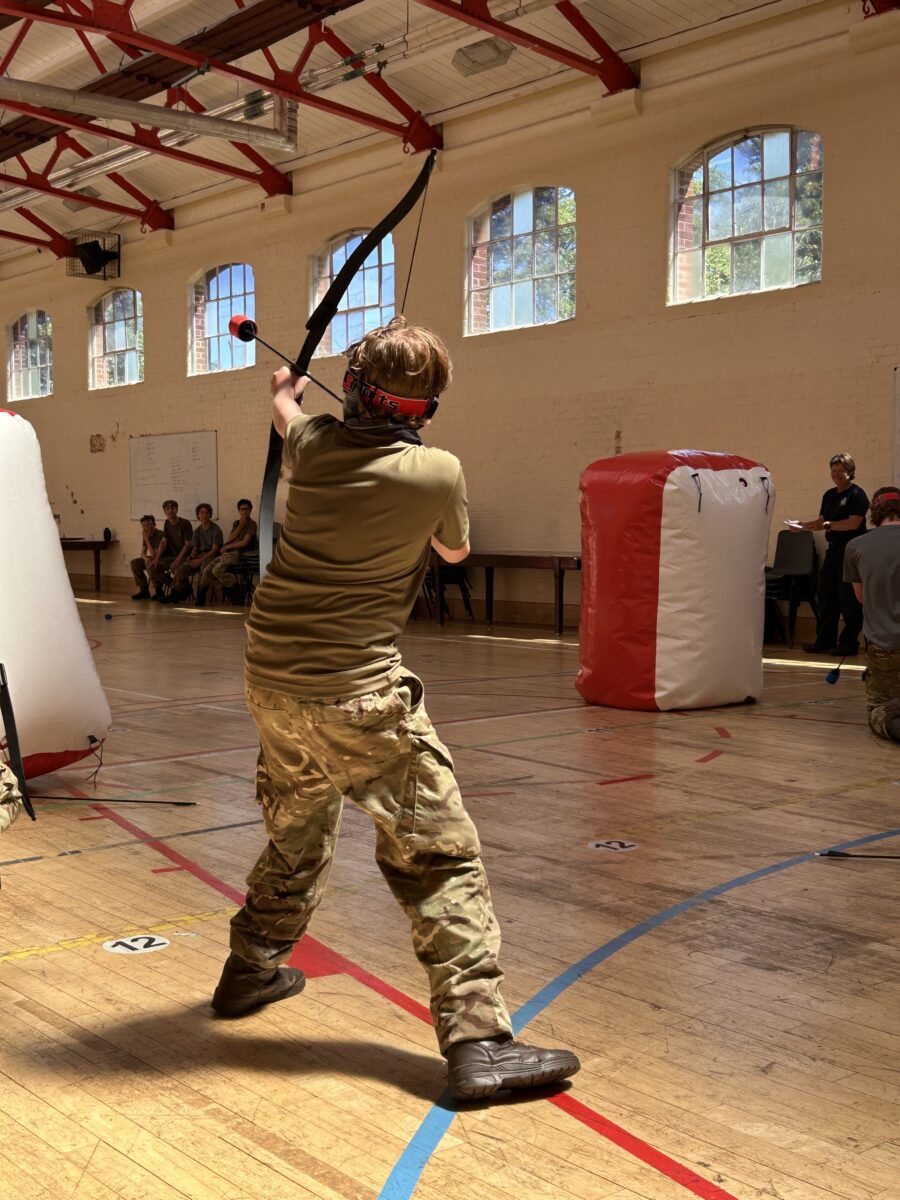 A person in camouflage clothing is actively engaged in archery indoors, aiming a bow and arrow towards a target that is not fully visible. There are inflatable obstacles in the room and other individuals in the background.