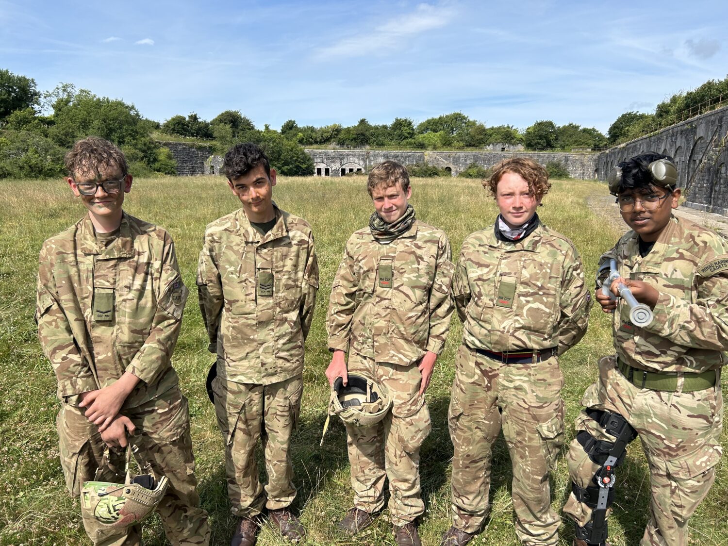 Five individuals dressed in camouflage fatigues are standing outdoors in a grassy field with some old stone structures in the background. They appear to be posing for a group photo.
