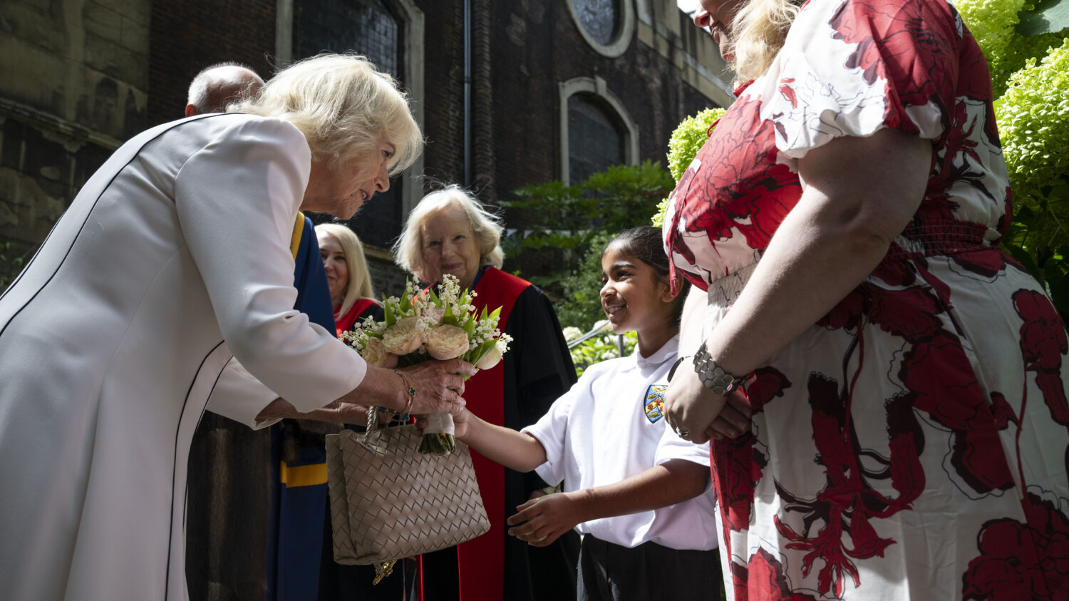 Queen Camilla, wearing a white jacket, is receiving a bouquet of flowers from a young girl. Another woman in a red and white patterned dress and a woman in academic regalia are also present. The scene appears to be outdoors, with a building and greenery in the background.