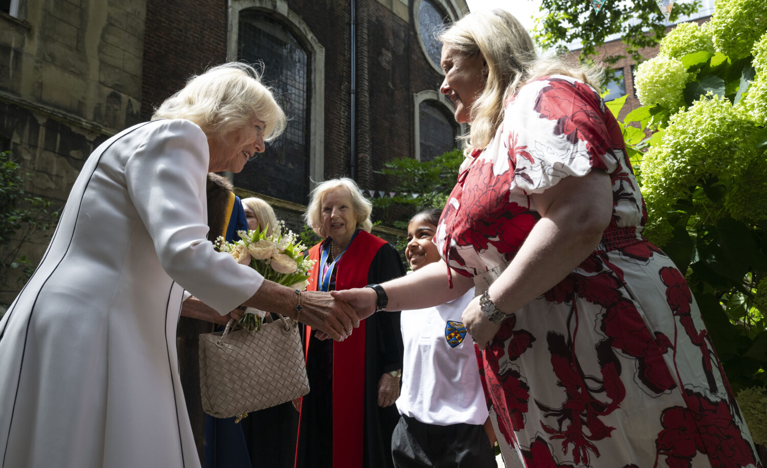 This image captures a moment during Queen Camilla's installation as an Honorary Freeman and Liveryman of the Worshipful Company of Stationers and Newspaper Makers on July 15, 2025. Queen Camilla, on the left, is seen shaking hands with a woman in a red and white floral dress. Queen Camilla is holding a small bouquet of light-colored flowers. In the background, to the right of Queen Camilla, an older woman in academic-style robes (red with blue trim) is visible, along with a younger person in a school uniform, both looking towards the interaction. The setting appears to be outdoors, with a building made of brick in the background to the left and green foliage on the right.