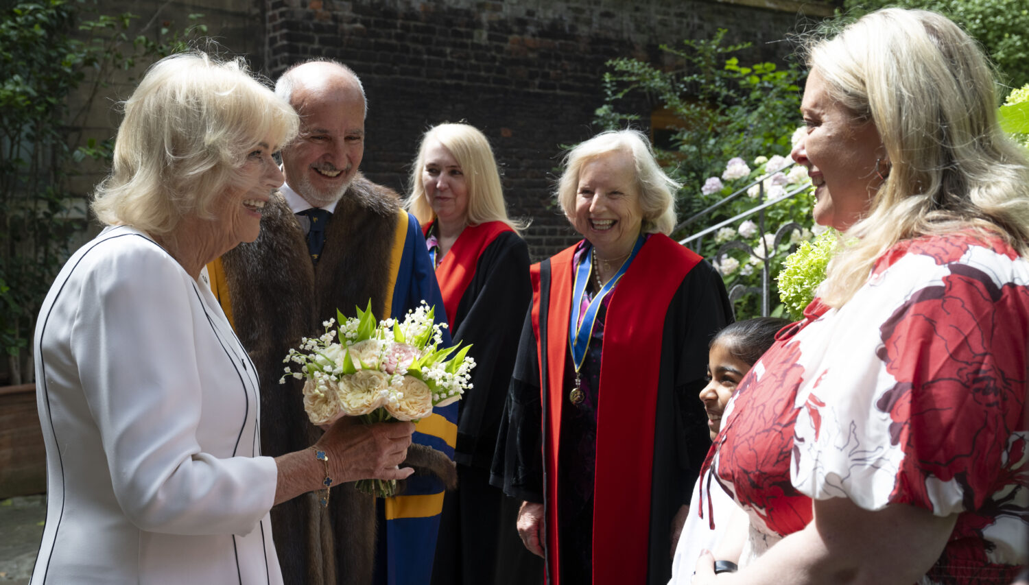 Queen Camilla, on the left, is looking towards and possibly speaking with the woman in the red and white floral dress, who is smiling. Queen Camilla is still holding the bouquet of flowers. Between Queen Camilla and the woman in the floral dress, a man in a fur-trimmed robe and a woman in red academic-style robes are visible, both smiling and looking towards the interaction. The young person in the school uniform is also visible on the far right, smiling and looking in the same direction. The background is similar to the first image, with a brick building and green foliage.