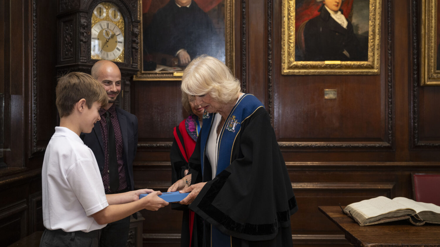 Queen Camilla, wearing academic regalia, is presented with a blue box from a young boy. Both are smiling. A man is standing behind the boy, also observing. The background features dark wood paneling, a grandfather clock, and framed portraits, suggesting an indoor setting similar to the previous image.