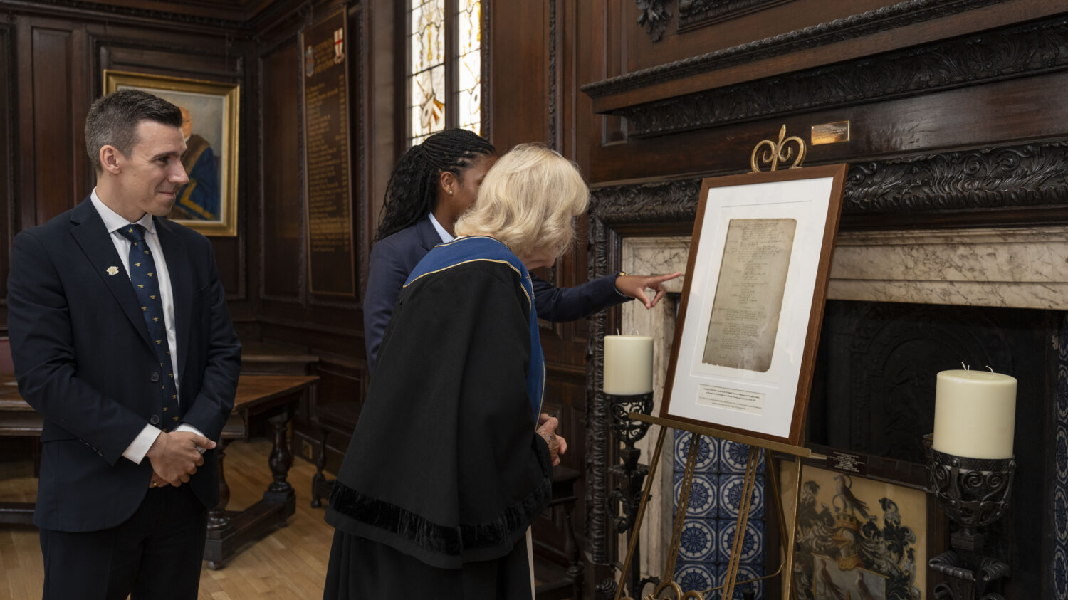 Queen Camilla, dressed in academic regalia, is looking at a framed document on an easel. Another person, possibly a student, is standing next to her and pointing at the document. A man in a suit is standing to the left, observing. The setting appears to be an ornate room, likely historical, with dark wood paneling and a fireplace.