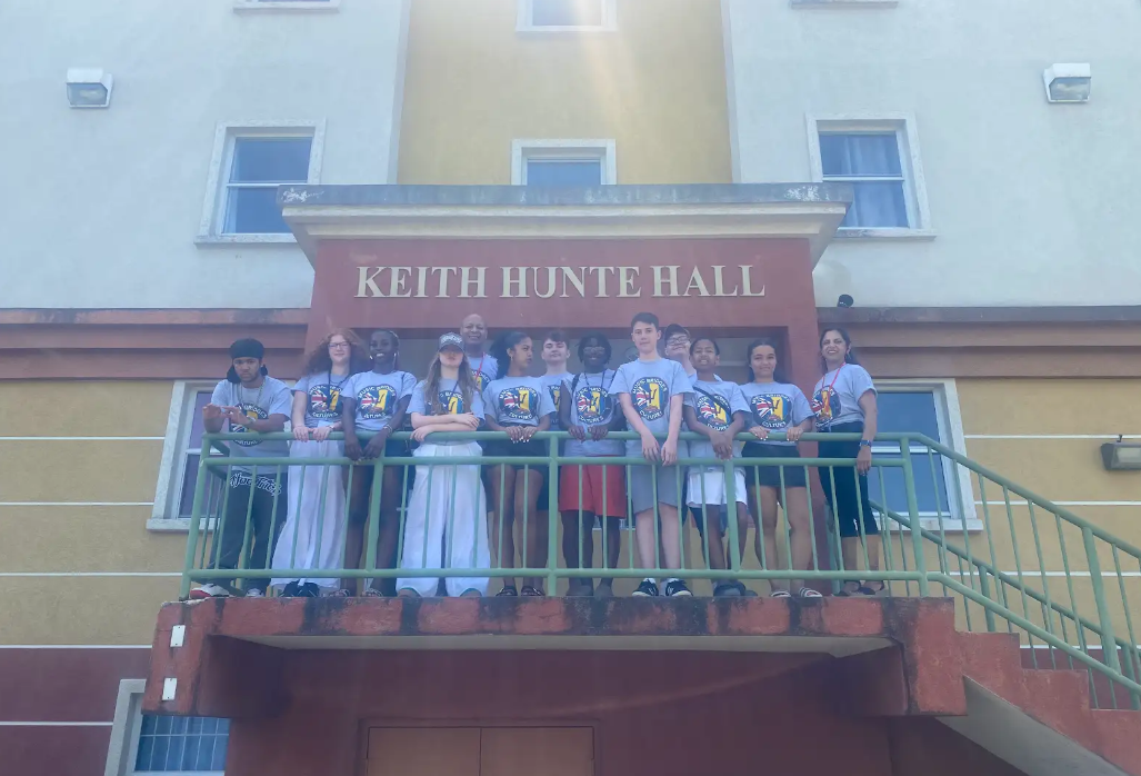 A group of young people and at least one adult are standing on the balcony/stairs of a building that has "KEITH HUNTE HALL" written above the entrance. They are all wearing similar grey t-shirts, suggesting they might be part of an organized group or event.