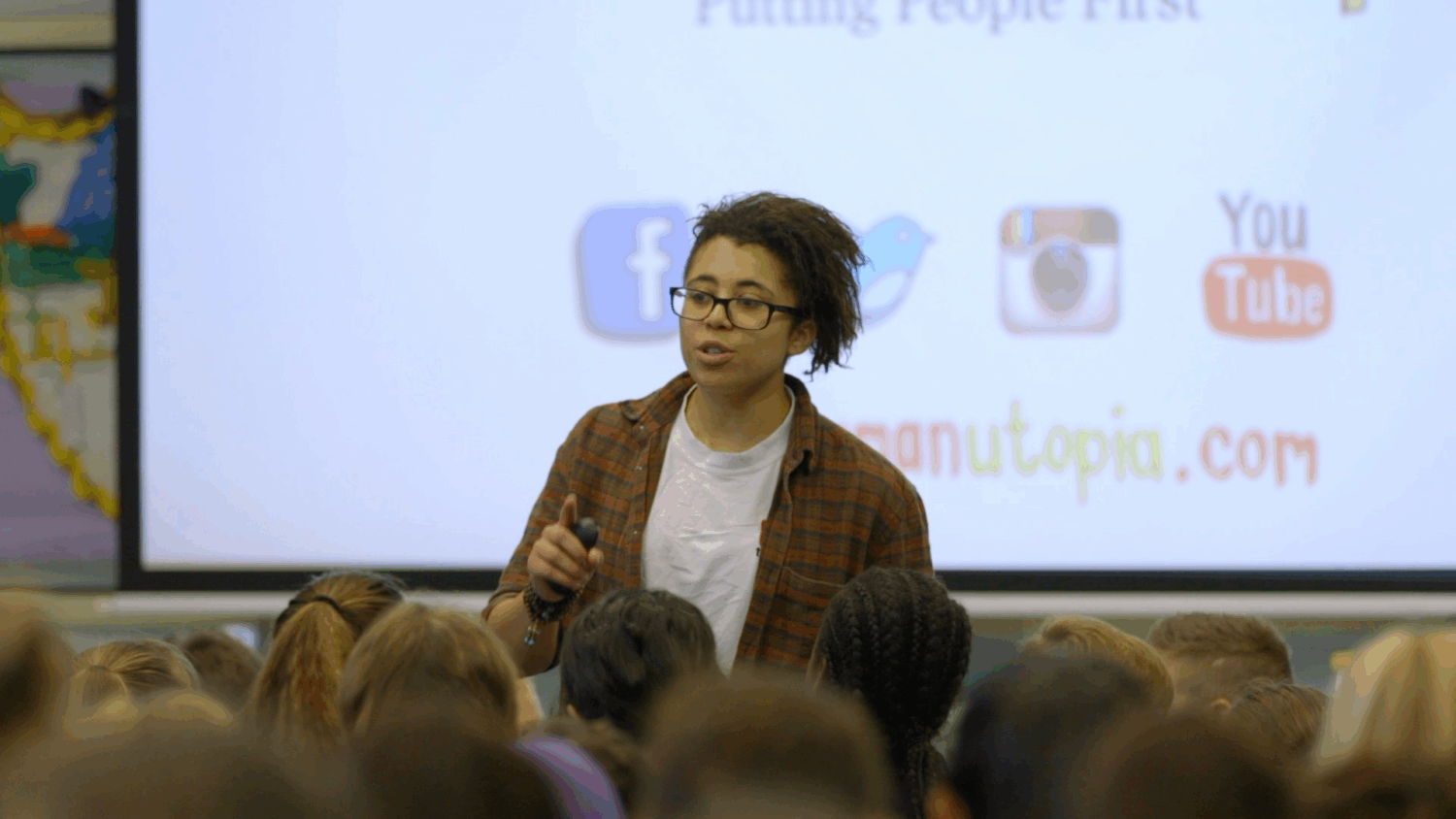 A person with glasses and a plaid shirt is speaking to a group of people, with their backs to the camera, in what appears to be a classroom or lecture hall. Behind the speaker, a presentation is being displayed on a screen with social media logos (Facebook, Twitter, Instagram, YouTube) and a website address.