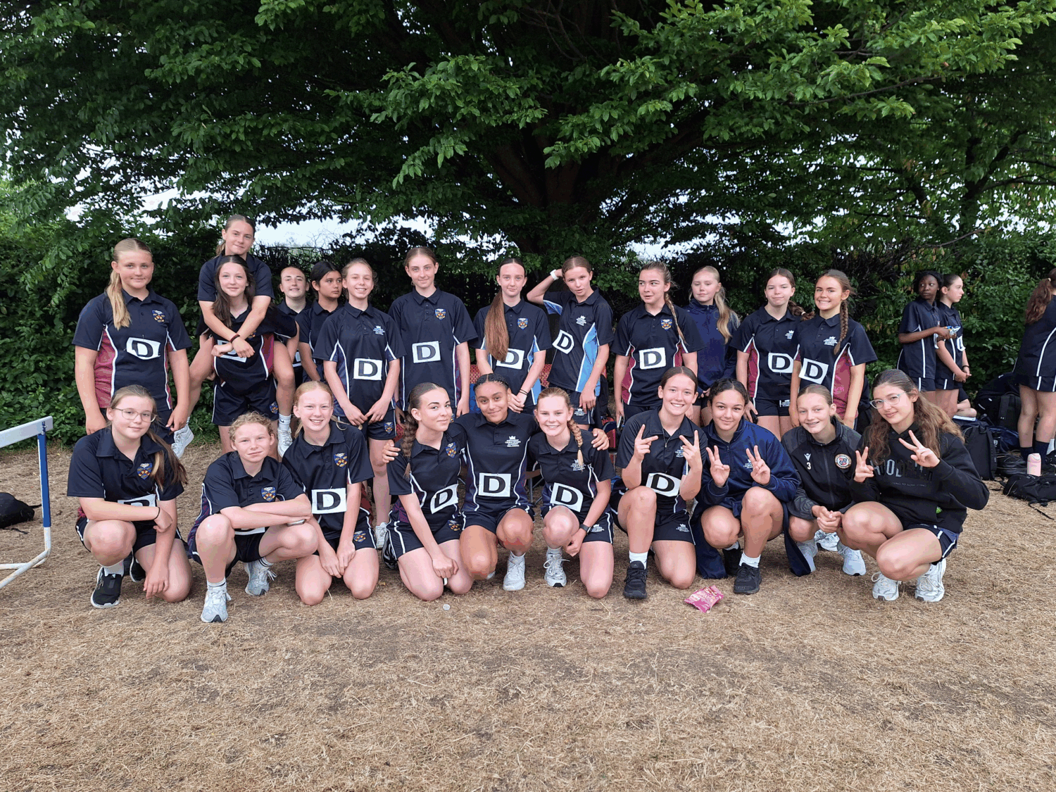 A large group of girls, likely a sports team, are posing for a group photo outdoors under a tree. Most of them are wearing matching dark-colored sports uniforms with a "D" logo, suggesting they are part of a school or club team. Some are standing, and others are kneeling or crouching in front.