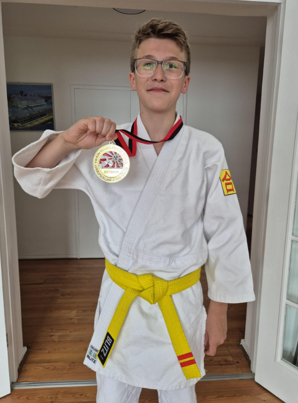 A young person wearing glasses and a white martial arts gi with a yellow belt is proudly holding up a gold medal. This suggests they have participated in and achieved success in a martial arts competition.