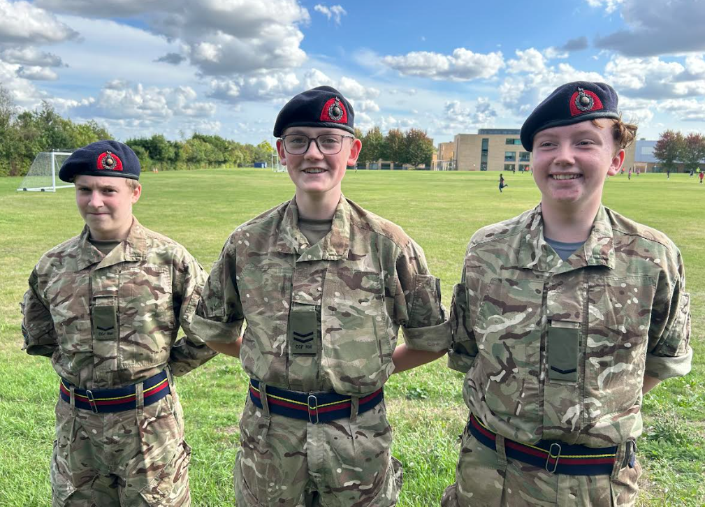 three young people are standing together in a grassy, open field, likely a school or college campus. They are wearing camouflage uniforms with insignia, including berets and belts. They appear to be part of an organization like the Army Cadet Force or a similar military youth group. The individual in the center is wearing glasses and all three are smiling, suggesting they are happy to be there. In the background, there are school buildings and other people can be seen on the field. The weather looks pleasant with a partly cloudy sky.