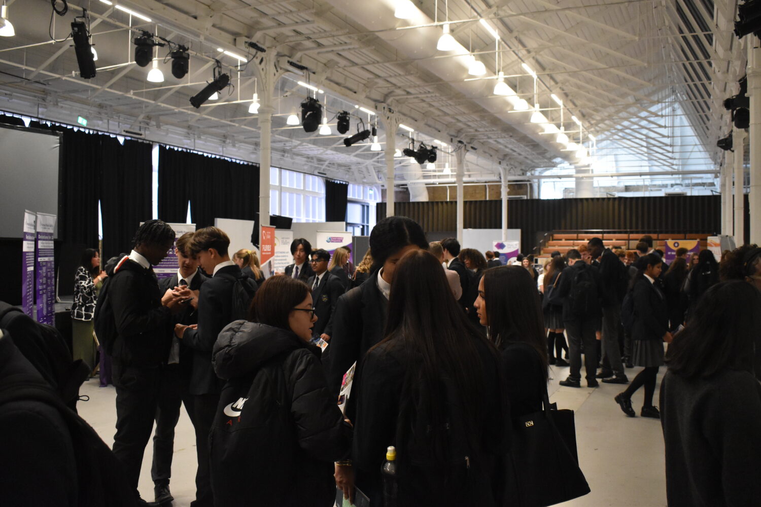 A group of students and adults are standing together, facing away from the camera, in a large, plain room. This could be a presentation or a talk, a common activity at careers fairs.