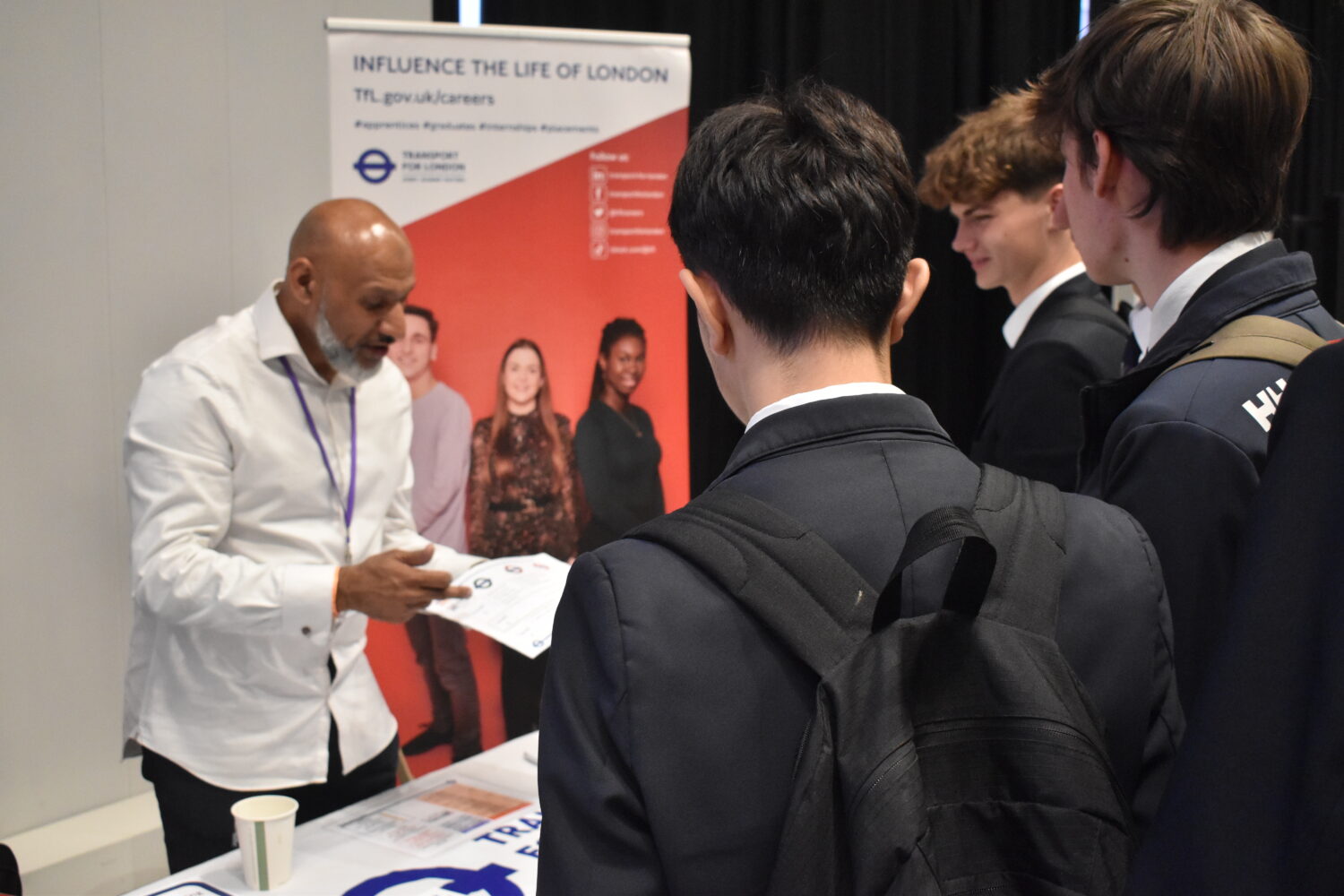 This picture shows a professional from "Transport for London" (TfL) talking to a group of students. He is pointing to a document on his table, likely explaining job opportunities or training programs. The "INFLUENCE THE LIFE OF LONDON" banner in the background clearly indicates a focus on careers within the city.