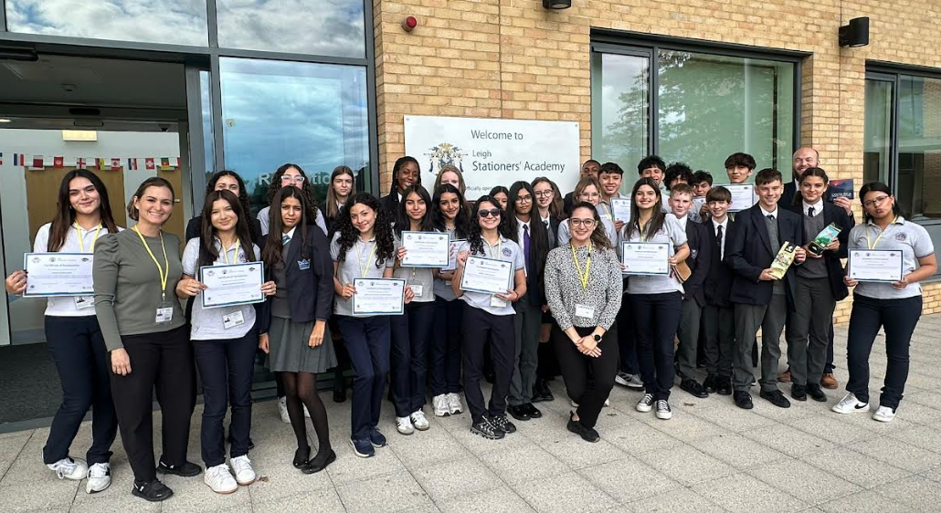 This image appears to show a group of students and teachers from Leigh Academy (the sign reads "Welcome to Leigh Stations Academy"). The people are standing outside the entrance of the building. Most of the students are holding certificates or documents, and a few are holding small gifts or packaged items (like bottles or boxes). This suggests they are participating in a group photo.
