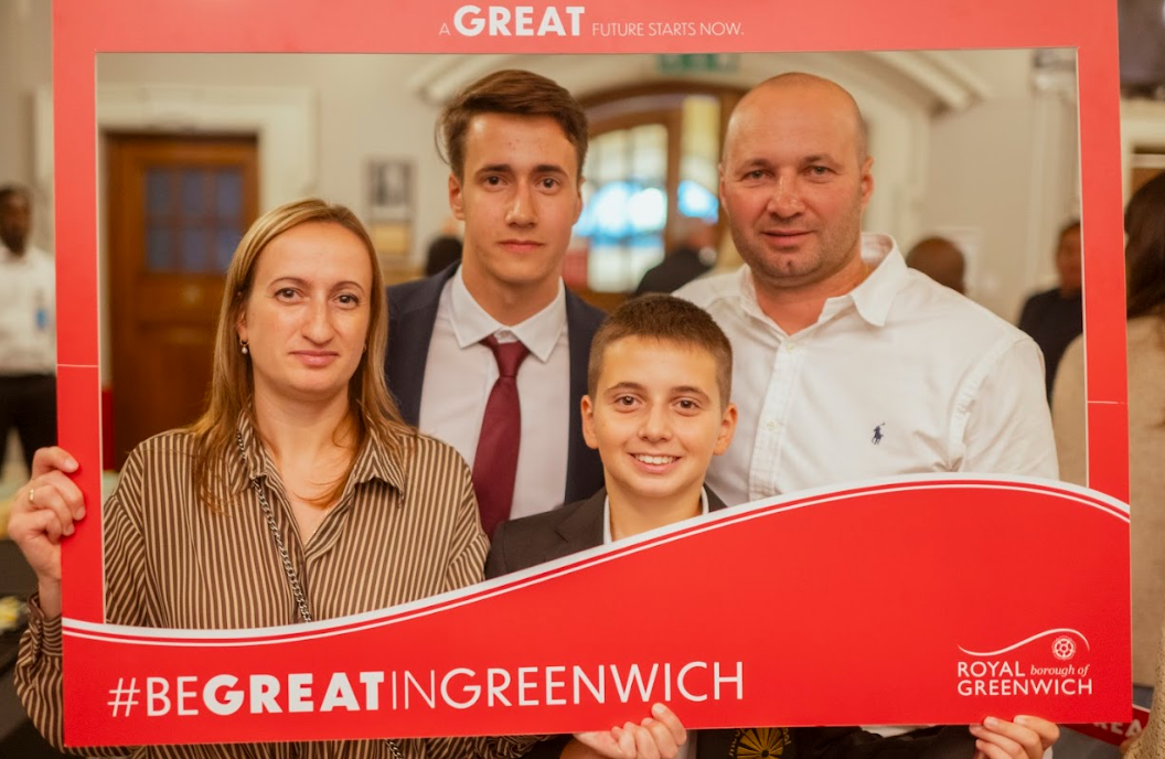 This is a family photo taken inside a large red frame. The text on the frame says "A GREAT FUTURE STARTS NOW" and "#BEGREATINGREENWICH" with the logo for the Royal Borough of Greenwich. This is likely a promotional photo booth at an event or ceremony hosted by the Royal Borough of Greenwich. The family—composed of two adults and two minors—is posing for the picture, supporting the message of the local area.
