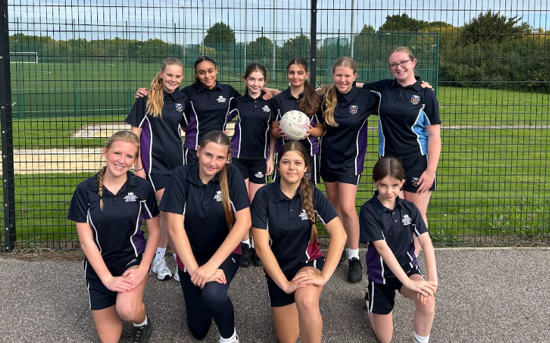 The photo itself is of a sports team, likely a netball team, standing on a sports pitch in front of a chain-link fence. There are eleven girls in matching black and purple sports uniforms. They are posing together as a team, with one of the girls holding a netball. This looks like a team photo following a game or practice session.