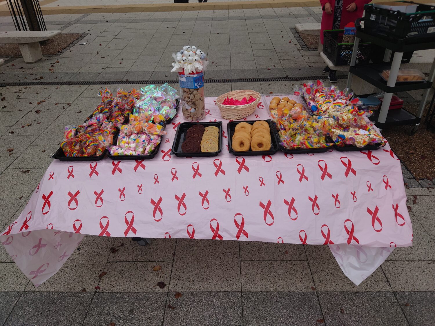 This image shows an outdoor stall set up on a paved area, likely for a fundraiser or market. The stall is selling a variety of treats and small novelty items. Items include many bags of colorful candies and sweets wrapped in cellophane. There are trays of donuts and cookies (chocolate chip and plain shortbread style). In the center, a large plastic jar holds several small plush toys (possibly dogs or snowmen) on sticks, possibly offered as prizes.