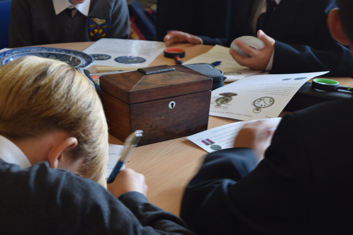 A close-up view showing students writing and looking at worksheets that feature images of items like a pocket watch and possibly medals or coins. A small wooden box is prominent on the table, and one person is holding a light-colored, smooth object.