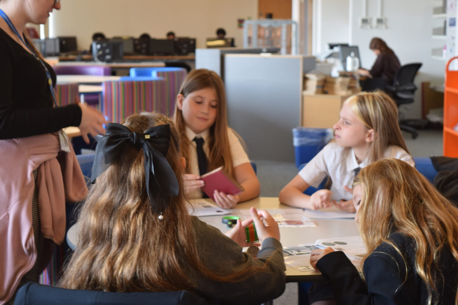 This image shows a group discussion setting. Four students are at a table, and an adult (possibly a teacher) is standing next to them. One student is holding a small notebook, while the others are engaged in conversation and looking at their papers. The background reveals the setting to be a bright, modern learning space with computer stations and bookshelves.