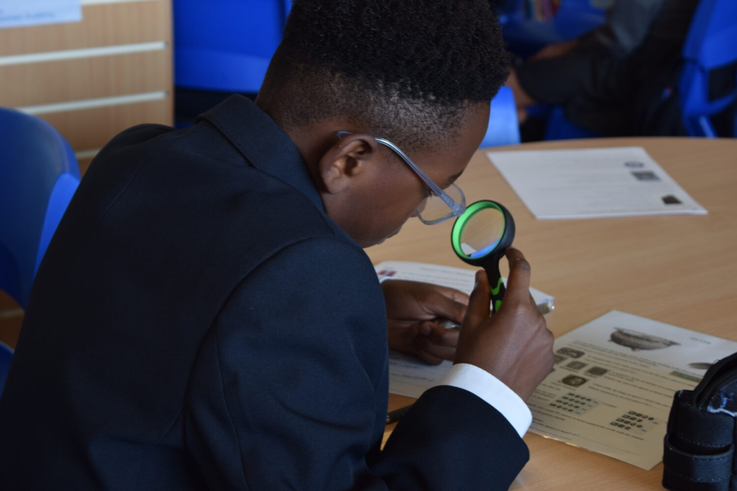 A student is using a magnifying glass to closely examine details on the documents or objects, indicating a task that requires close observation.