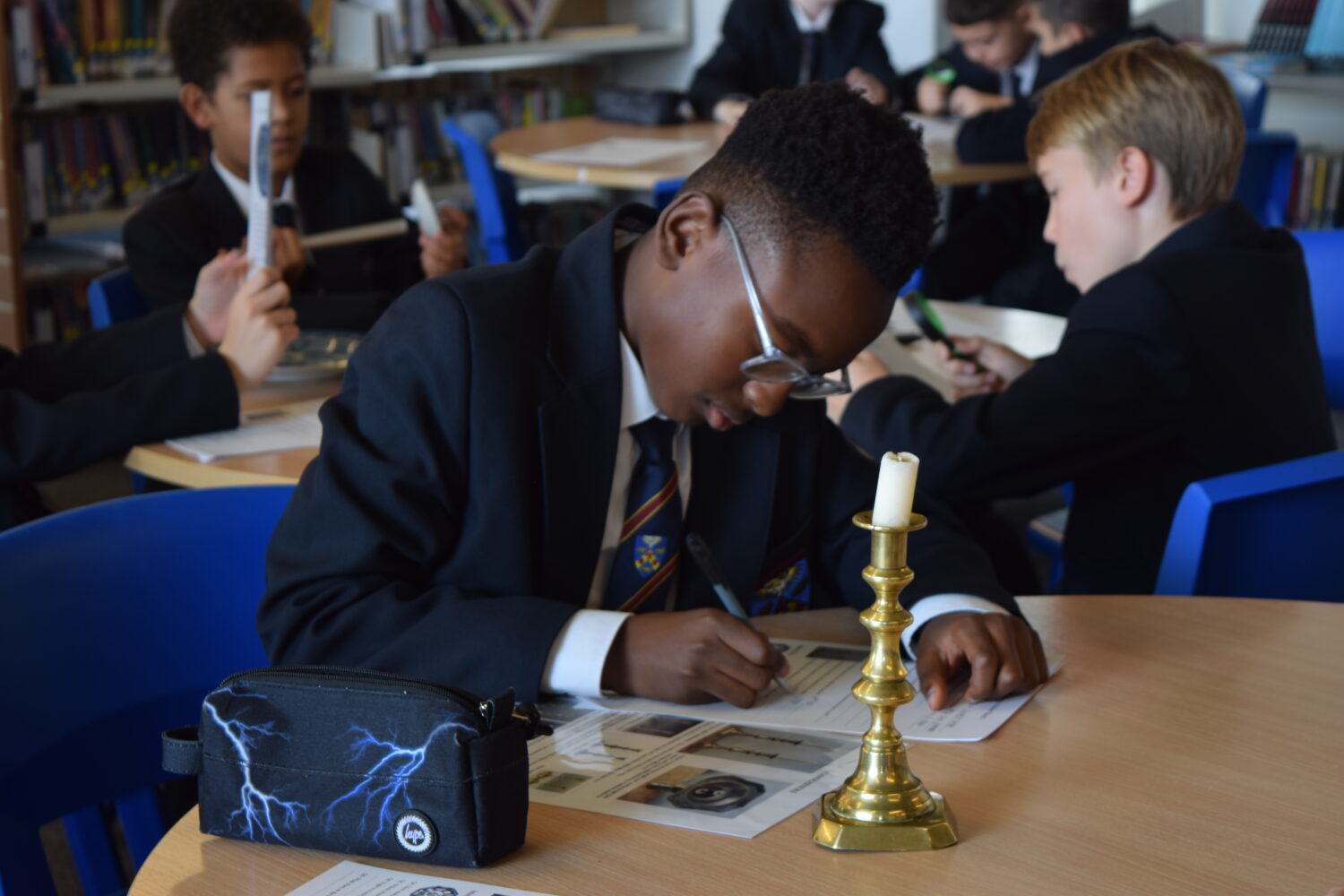 A student is deeply focused on the activity. he is writing on a worksheet with a brass candlestick and unlit candle nearby, suggesting a historical or 'old-fashioned' theme to the items they are studying.