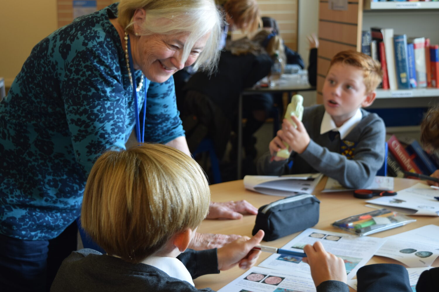 An adult, possibly a teacher or presenter, is interacting directly with the students. She is smiling and speaking to the students, one of whom is holding the artifact and talking excitedly.