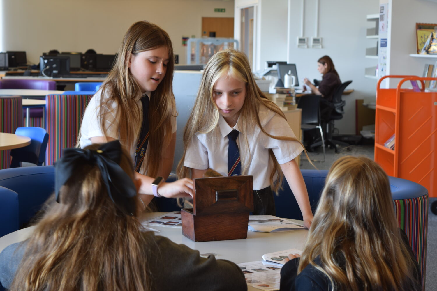 Four students, wearing school uniform shirts and ties, are gathered around a table. Two girls are focused on a small wooden box that is open, perhaps examining its contents or an item placed nearby. They also have worksheets spread out on the table.