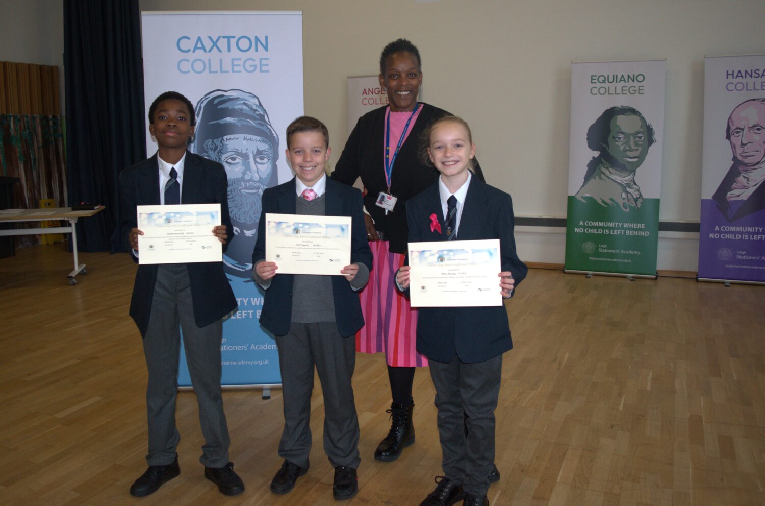 This image shows three students smiling and proudly holding their certificates, posing with a female staff member who is standing slightly behind them. The college banners for Caxton, Angelou, Equiano, and Hansard are visible in the background.