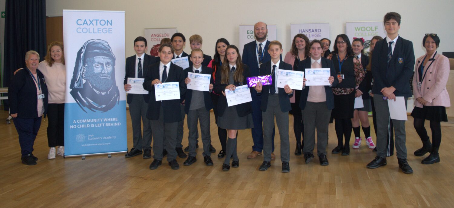 This is a group photo of multiple students, all holding their certificates (one student is also holding a small gift, possibly a chocolate bar), posing with a larger group of staff members. The staff are positioned both at the front and behind the students. The college banners are once again visible behind them.