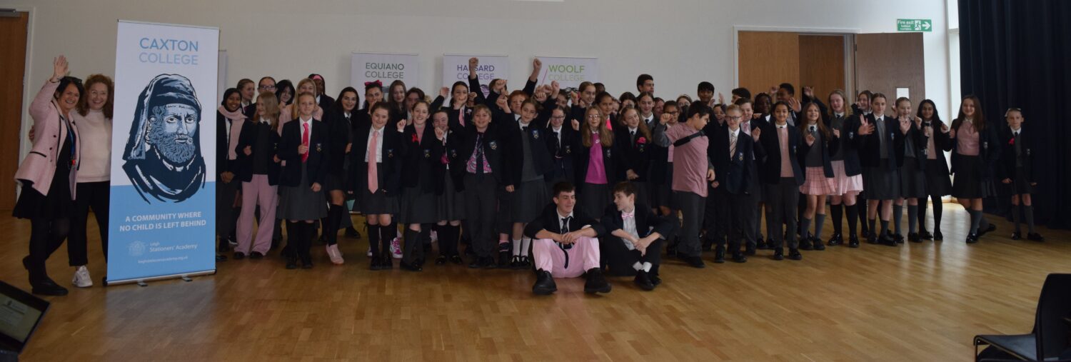 A large group of students in school uniform are gathered together, posing enthusiastically with their hands raised. Many wearing an item of pink clothing