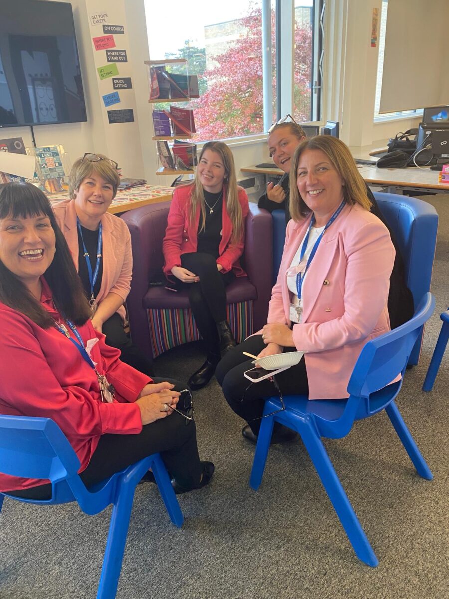 A group of five female staff members are gathered in a brightly lit, casual seating area, which looks like a quiet reading or breakout corner in the school. They are sitting in colorful blue and purple chairs and smiling at the camera, engaged in a conversation or break. Three of the five are wearing bright pink jackets or tops.