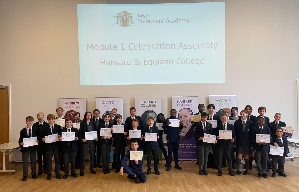 This is a large group photo of students and some staff members taken during the "Module 1 Celebration Assembly." The students are dressed in school uniform and are standing in rows, proudly holding certificates. A projection screen on the wall confirms the event's name and features the academy's logo and the specific colleges being celebrated: Hansard & Equiano College. The banners for all five colleges (Angelou, Caxton, Equiano, Hansard, and Woolf) are visible in the background.