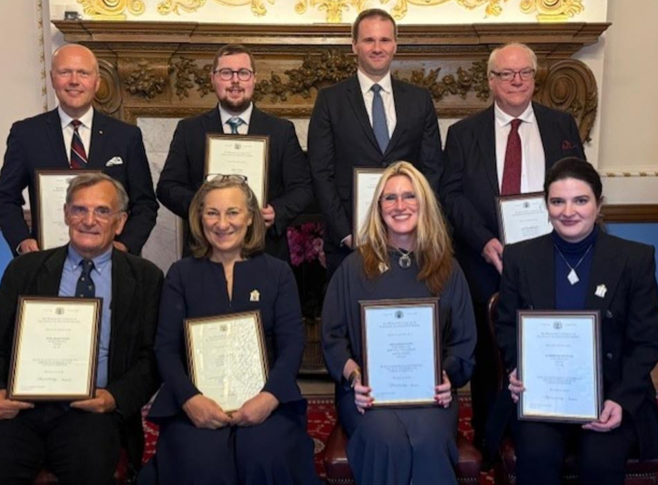 Eight formally dressed award recipients are posing for a photograph. They are standing and sitting in a traditional, ornate room in front of a decorative fireplace, each holding a framed certificate that recognizes their achievement or fellowship.