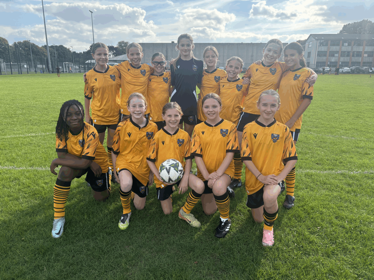 This photograph shows a girls' soccer (football) team posing for a team picture outdoors on a sunny day