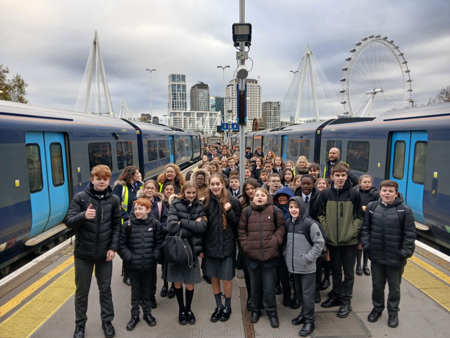 A large group of students and a few adults are gathered on a railway platform between two modern blue and light blue trains. The group is smiling and posing for the camera, with the young people generally wearing dark coats and some appearing to be in school uniforms (dark skirts/trousers). The background clearly identifies the location as a city center station, likely in London, as the skyline features modern skyscrapers, the distinctive white suspension cables of the Golden Jubilee Bridges, and the London Eye Ferris wheel. The sky is overcast.
