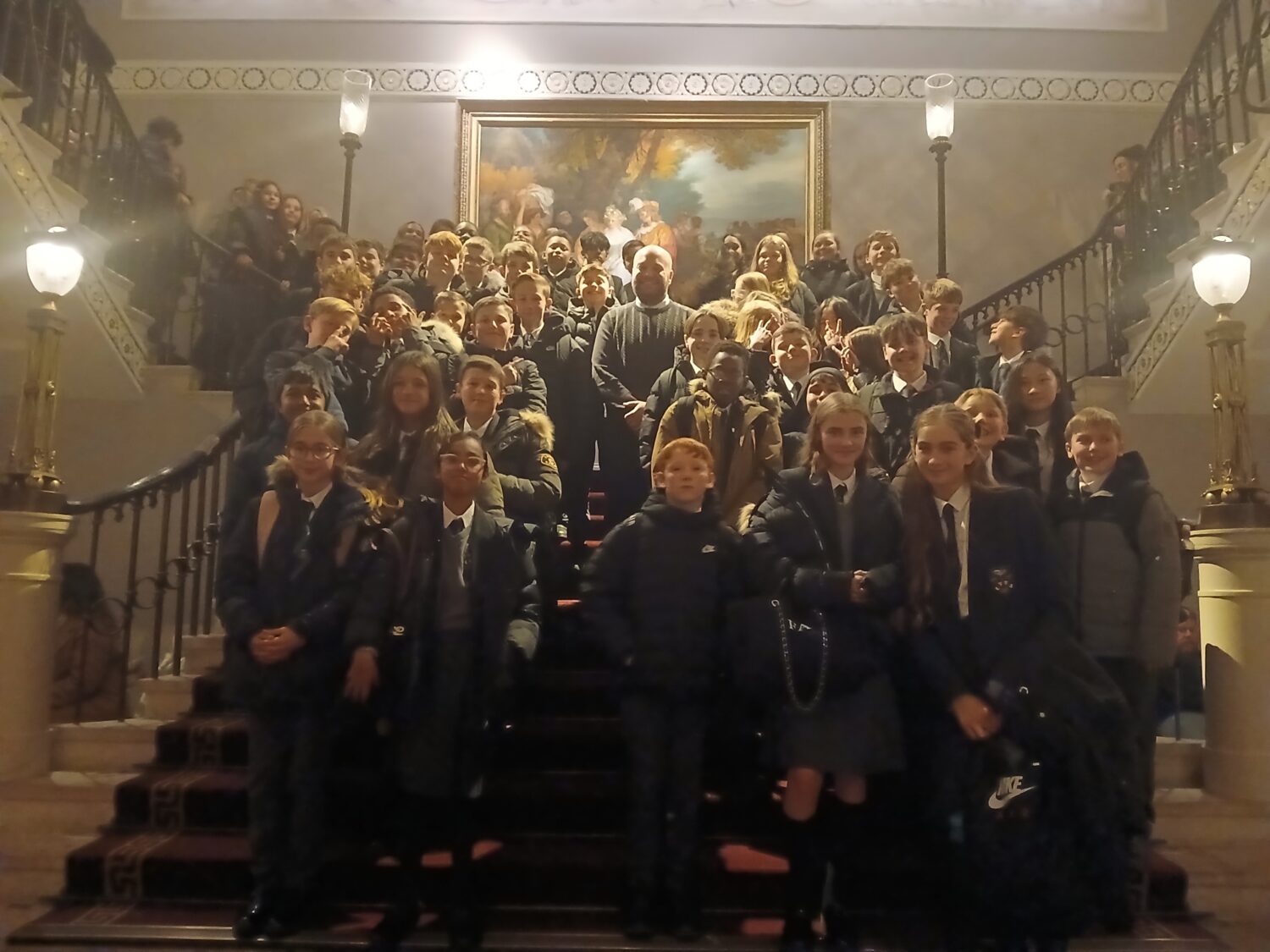 A large group of students and a few adults are posing for a photo on a grand, ornate staircase inside a building. The stairs are carpeted in red and flanked by stone balustrades topped with large, classical lamps. The group is spread across the width and height of the staircase. Most of the young people are wearing dark coats and what appears to be a school uniform (dark blazers and ties/white shirts). An adult, possibly a teacher or tour guide, stands near the center of the group, slightly elevated. Above the group on the wall is a large, classic oil painting in a gold frame. The interior is well-lit but with a warm, indoor glow.